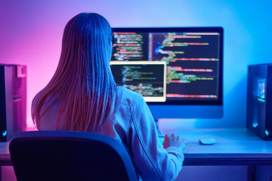 Persona escribiendo c&oacute;digo en una computadora en una habitaci&oacute;n estilo ne&oacute;n, vista desde atr&aacute;s