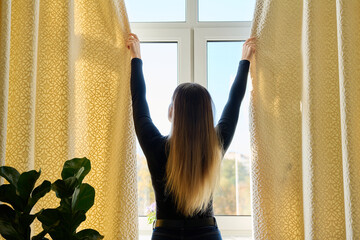 Young woman closing opening thick curtains on window
