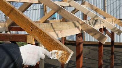 A painter in dirty clothes and gloves uses a brush to apply orange paint or varnish to a wooden structure in the street against a lumber fence.