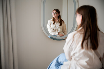 Beautiful girl looking away in mirror and smiling. Pretty female checking herself. Cute girl sitting in chair opposite to mirror at home.