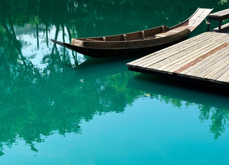 Obraz premium A beautiful Asian-style wooden boat and wooden dock are in the turquoise lake under the morning sunlight. Trees reflect on the water's surface, and there is space for text.