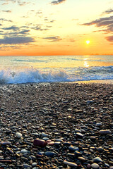 Stone beach on sunrise. Sunset at sea, view from shoreline of pebbles beach. Sea beach shore with waves in ocean. Shore landscape on Spain resort. Ocean shoreline scenic. Waves in sea. Vertical photo.