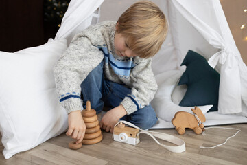 6-year-old boy sits in a wigwam tent, playing with wooden toys with interest. Children's room, childhood, games, imagination © Anna