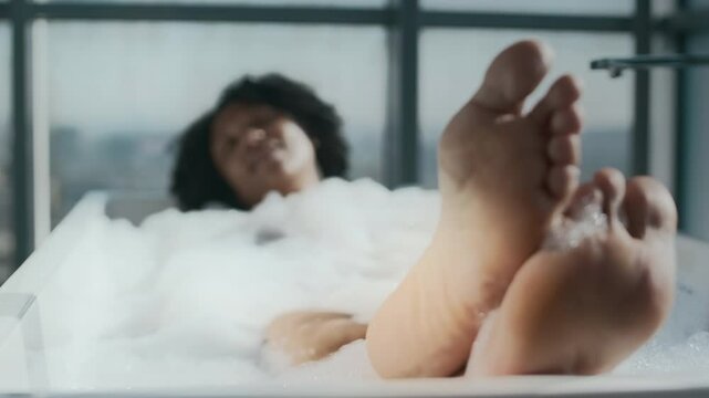 Delighted young Black woman relaxing in comfortable freestanding bathtub filled with soapy warm water, with natural sunlight coming from panoramic windows. Selective focus on woman feet in foreground
