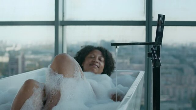 Tranquil young Black woman having bath with stunning city view in high-rise bathroom, lying in soapy water with her eyes closed, creating atmosphere of luxury and indulgence