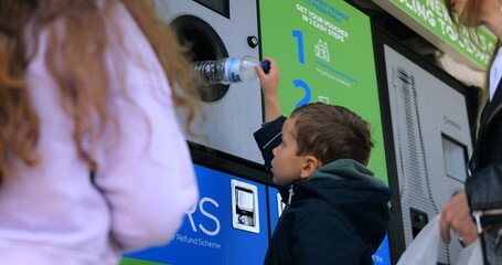 Toddler boy puts used plastic bottle into reverse vending machine on city street. Little child...