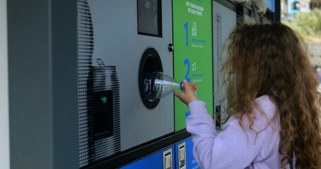 Little girl with long curly hair puts plastic bottle into reverse vending machine on city street. Teaching children to sort garbage everyday
