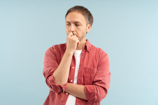 Worried man stands with tense posture, deep in anxious thoughts, nervously biting nails looking downcast as wrestles with inner stress. Distressed male closed off absorbed feels anxious, withdrawn.