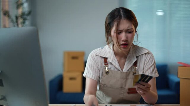 Frustration builds as an asian woman entrepreneur counts money at her desk, facing challenges in her small business