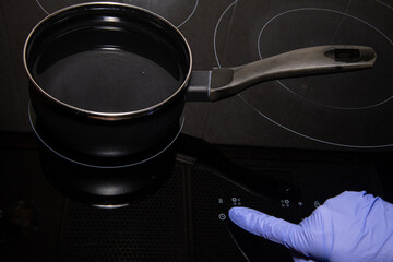 Gloved finger on the touch panel of a ceramic hob bringing a pan with water to the boil