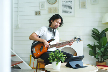 Attractive Japanese Man practicing a bass guitar with online learning on digital tablet while...