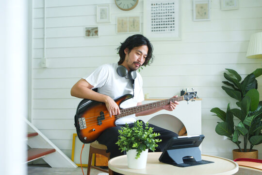 Attractive Japanese Man practicing a bass guitar with online learning on digital tablet while wearing headphones in Living Room. - Powered by Adobe