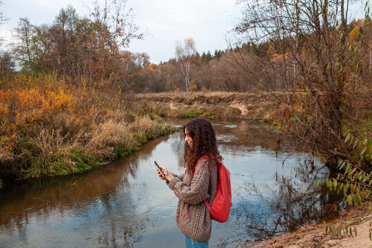 Side view of curly woman with backpack standing on river bank looking at phohe screen. Traveler building route in wildlife national park. Concept of ecotourism, hiking, travel in autumn.