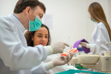 Latin female patients listen to a friendly dentist doctor who shows them how to brush their teeth using a jaw model in a dental clinic.
