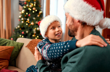 Dad and son are sitting together in a cozy, Christmas-decorated living room at home. The boy, wearing a Christmas sweater, hugs his dad. It’s a special time together on Christmas.