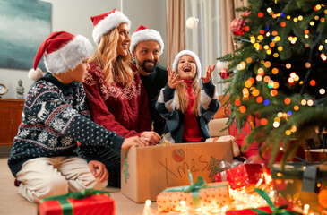 A happy family is preparing for Christmas and New Year. Cheerful joyful girl throws up a Christmas tree decoration. Under the tree there is a box with Christmas decor and gifts.