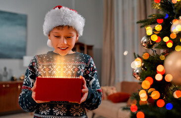 A happy smiling boy in festive Christmas clothes is standing near a Christmas tree and holding a red gift box from which light is radiating. Christmas gift.