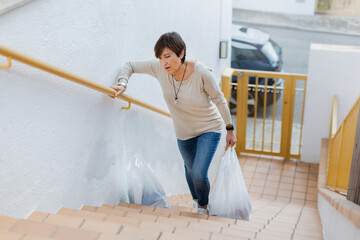 A determined Senior woman tirelessly struggles to carry heavy grocery bags upstairs in her busy home daily work. Daytime, outdoors. Horizontal