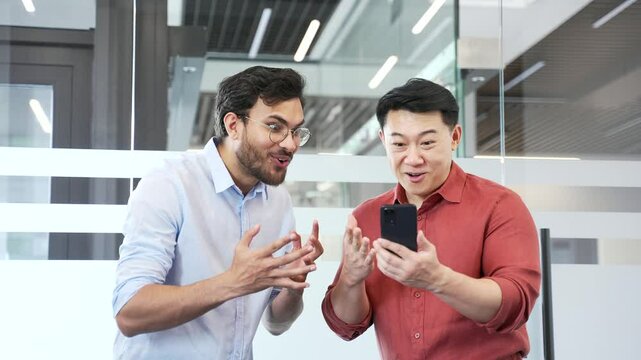 Two excited businessmen watching sports match or cheering for bids at the auction using mobile phone in business office. Happy colleagues celebrating success showing excitement and achievement