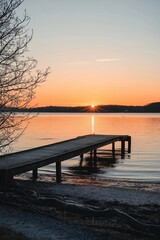 Obraz premium A wooden pier stretches into a calm lake at sunset, surrounded by a tranquil, scenic landscape and colorful sky.