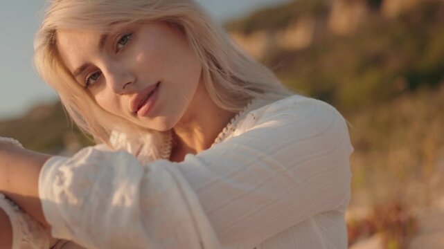 Close up of a beautiful young blonde woman with green eyes and a white dress looking at the camera and smiling on a warm summer evening during golden hour.