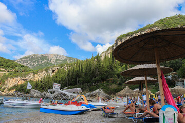 Beach View with Straw Umbrellas, Tourist Boats, and the Sea with Hilly Landscape of a Greek Island