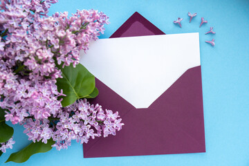Composition with empty purple envelope and beautiful spring lilac flowers on blue background. Mockup card invitation greeting card postcard copy space template blank. Branches of lilac blooming