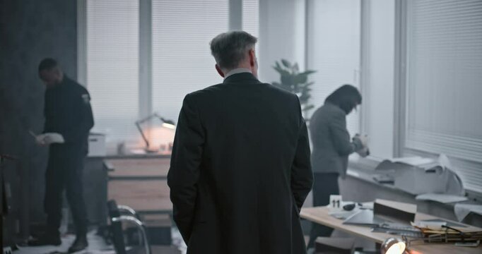 Office robbery case: Back view of male detective in suit examining crime scene. Female forensic scientist takes photos of clues and evidence, African American police officer searches fingerprints.