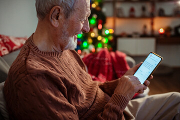 Senior man texting on smartphone by Christmas tree at home