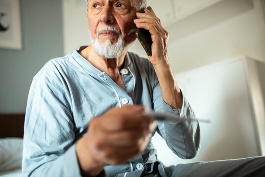Senior man holding thermometer making phone call at home