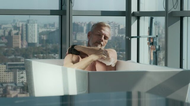 Handsome Caucasian adult man with gray hair and beard washing himself with bath sponge while sitting in freestanding bathtub filled with soapy water, set up against floor-to-ceiling window
