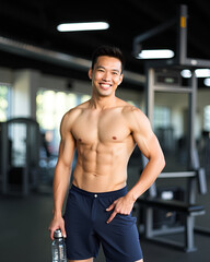 asian man is posing in the gym with a cheerful expression and carrying a bottle