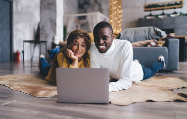 Happy couple lying on the floor, smiling while browsing a laptop together in a cozy, holiday-decorated home. They enjoy online Christmas shopping, sharing festive joy and warmth