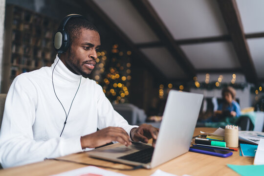 African American man working remotely on laptop, wearing headphones and focused. Cozy workspace with warm lighting, festive decorations. Background shows relaxed atmosphere and blurred holiday lights