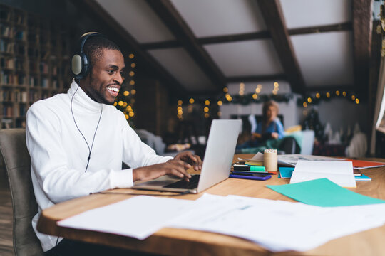 African American man smiling while studying IT, wearing headphones, focused on laptop in cozy, festive home setting. Background shows warm holiday lights and a relaxed ambiance, remote learning