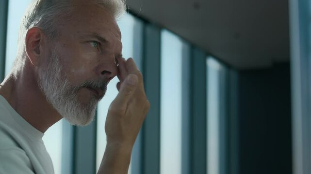 Caucasian adult man in white T-shirt applying facial cream with concentration. Soft light creating inviting atmosphere, highlighting his dedication to skincare in stylish, modern setting