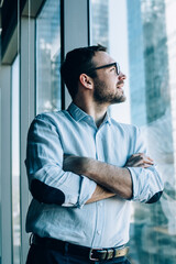 Contemplative male entrepreneur with crossed hands standing near office window view and feeling pondering during work day in company, Caucasian pensive corporate boss thoughtful looking away