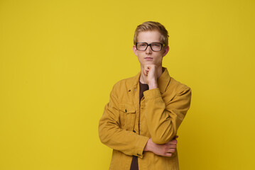 A young man with blond hair and glasses ponders a question, his hand resting thoughtfully on his chin. The bright yellow backdrop accentuates his serious expression