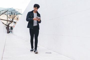 Fototapeta premium Trendy joyful ethnic man using tablet while walking along urban street tunnel