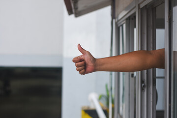 arm extending out of a window, with the hand giving a thumbs-up gesture. The background includes parts of a building and some blurred elements, possibly indicating an urban or residential setting.
