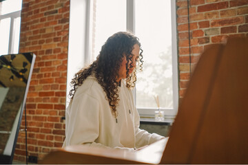 A woman with curly hair sits at a piano, enjoying the music she creates in a well-lit living room featuring cozy furniture.