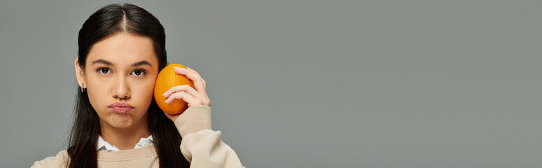 A young woman in fashionable attire holds an orange to her cheek, lost in thought and emotion.