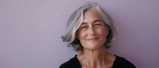 A woman with silver hair smiles gently against a muted purple backdrop, embracing elegance and confidence in a simple, contemporary setting.