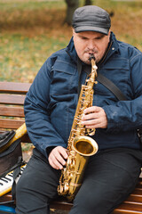 A saxophonist performs in a serene park during autumn while seated on a bench surrounded by fallen...