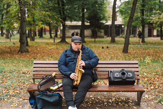 A saxophonist performs in a serene park during autumn while seated on a bench surrounded by fallen leaves
