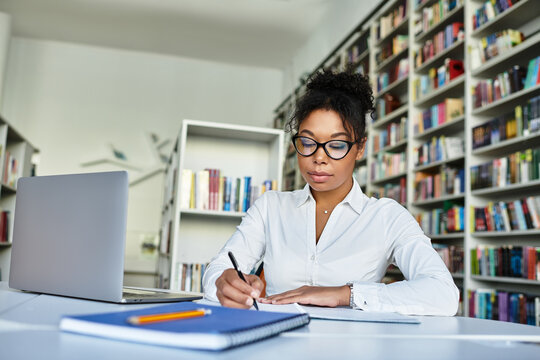 A dedicated African American teacher outlines plans at a desk surrounded by bookshelves.