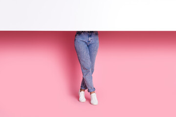 Naklejka premium Cropped photo of anonymous woman in denim jeans and standing tiptoes wearing sneakers hiding behind whiteboard isolated on pink color background