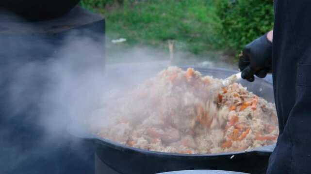 Chef in black mixing steaming plov in a cauldron outdoors, showcasing the art of cooking in slow motion
