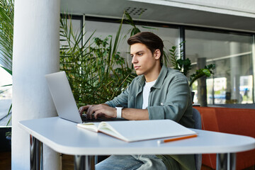 Concentrated individual engages with his laptop at a library table, surrounded by greenery.