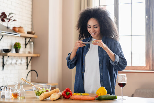 Happy Black food blogger girl taking picture of salad ingredients on mobile phones, recording video, shooting cooking process, reading recipe on Internet, chatting online in home kitchen